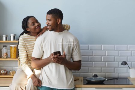 Foto de Pareja afroamericana sonriendo y hablando entre sí mientras usan un teléfono inteligente, pasan tiempo juntos en casa - Imagen libre de derechos