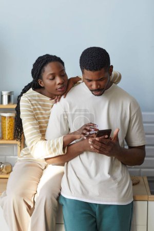 Téléchargez les photos : Couple afro-américain discutant message sur smartphone tout en se tenant dans la cuisine à la maison - en image libre de droit