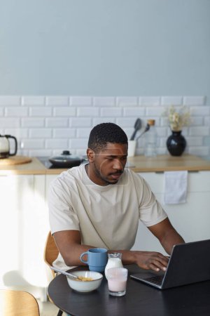 Téléchargez les photos : Afro-Américain travaillant sur ordinateur portable pendant le petit déjeuner assis à table dans la cuisine - en image libre de droit