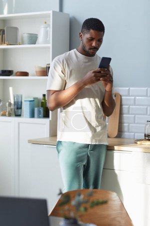 Photo for Vertical image of African American man reading message on smartphone while standing in the kitchen - Royalty Free Image