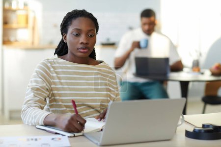 Téléchargez les photos : Afro-américaine prendre des notes dans un carnet tout en travaillant en ligne sur ordinateur portable assis à la table à la maison - en image libre de droit