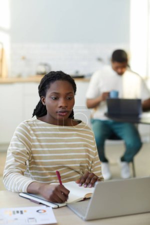 Téléchargez les photos : Afro-américaine jeune femme étudiant en ligne sur ordinateur portable assis à la table dans la cuisine à la maison - en image libre de droit