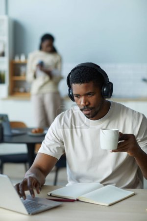 Téléchargez les photos : Afro-Américain dans les écouteurs boire du café le matin tout en travaillant en ligne sur ordinateur portable - en image libre de droit