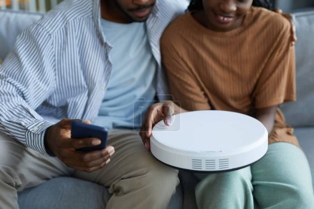 Téléchargez les photos : Gros plan du couple afro-américain reliant l'aspirateur robot au smartphone avant les travaux ménagers - en image libre de droit