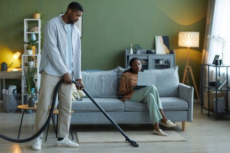 Téléchargez les photos : Homme aspirant le sol avec aspirateur dans le salon, il aide sa femme à faire des travaux ménagers - en image libre de droit