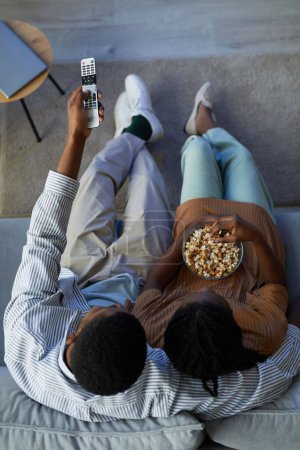 Téléchargez les photos : Image verticale du couple afro-américain regardant un film avec du pop-corn assis sur le canapé dans le salon - en image libre de droit