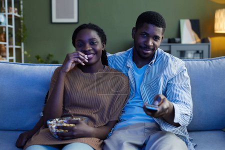 Foto de Afro americano feliz pareja disfrutando viendo película juntos en sofá y comer palomitas de maíz en casa - Imagen libre de derechos