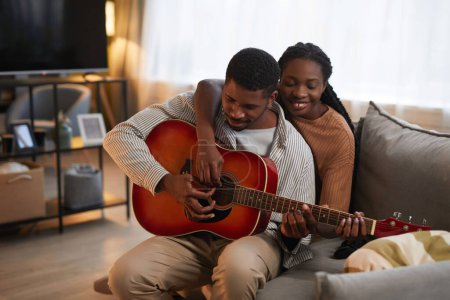 Téléchargez les photos : Heureux couple afro-américain apprenant à jouer de la guitare ensemble assis sur le canapé dans le salon - en image libre de droit
