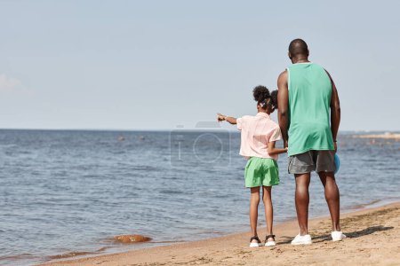 Téléchargez les photos : Vue arrière de papa marchant avec sa fille sur la plage - en image libre de droit