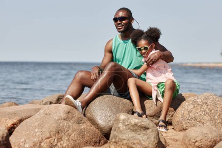 Photo for African American dad sitting on stones on coastline together with his daughter and they enjoying summer days - Royalty Free Image