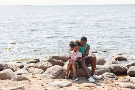 Téléchargez les photos : Afro-américain papa regarder vidéo sur smartphone avec sa fille alors qu'ils sont assis sur des pierres sur la plage - en image libre de droit