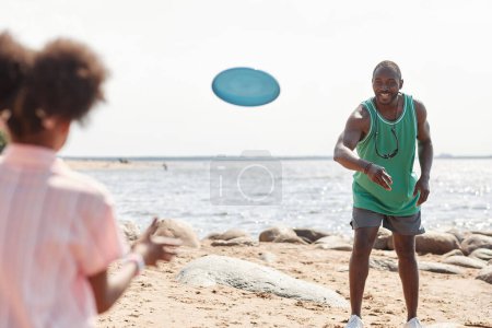 Foto de Feliz papá afroamericano jugando frisbee con un niño en la playa durante sus vacaciones de verano - Imagen libre de derechos