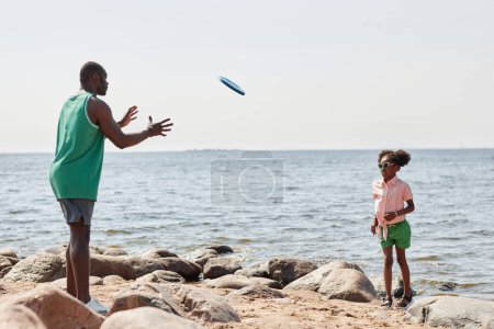Photo for African American dad throwing plate to his little daughter, they playing outdoor game on the beach - Royalty Free Image