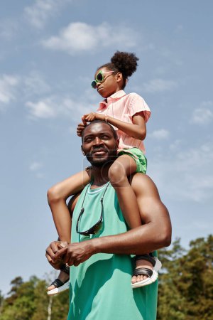 Téléchargez les photos : Image verticale du père afro-américain marchant avec sa petite fille à l'extérieur en journée d'été - en image libre de droit
