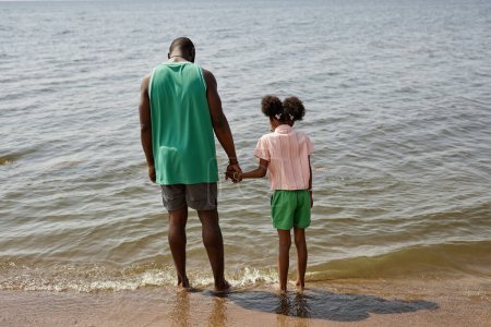 Téléchargez les photos : Vue arrière du père afro-américain et du petit enfant debout sur la plage et touchant l'eau avec les jambes - en image libre de droit