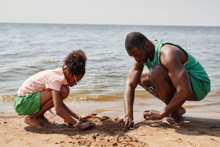 Téléchargez les photos : Afro-américain papa construire château de sable avec sa petite fille pendant qu'ils passent du temps sur la plage - en image libre de droit