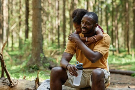 Téléchargez les photos : Papa boire de l'eau et se reposer avec l'enfant sur le rondin après la randonnée dans la forêt - en image libre de droit