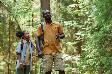 Téléchargez les photos : Papa afro-américain montrant sa fille nature sauvage lors de leur randonnée dans la forêt - en image libre de droit