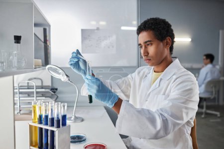 Photo for Side view portrait of ethnic young man holding test tube while doing research in laboratory, copy space - Royalty Free Image