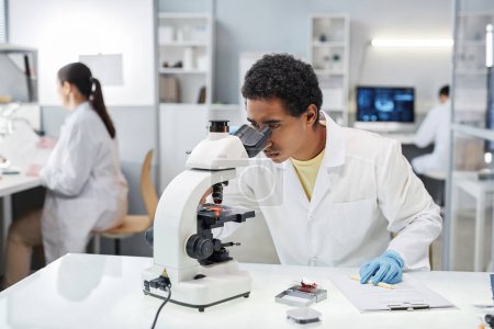 Téléchargez les photos : Portrait de jeune homme ethnique en tant que scientifique regardant au microscope tout en faisant des recherches en laboratoire, l'espace de copie - en image libre de droit