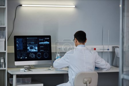 Photo for Back view at male scientist at desk in laboratory with data on computer screen, copy space - Royalty Free Image