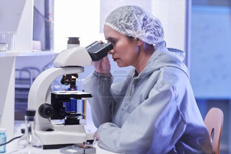 Téléchargez les photos : Portrait latéral d'une scientifique regardant au microscope sous la lumière du néon tout en faisant des recherches en laboratoire - en image libre de droit