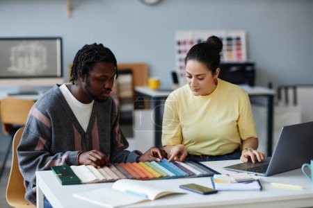 Téléchargez les photos : Jeunes collègues discutant des couleurs sur la palette tout en travaillant ensemble en équipe debout à la table au bureau - en image libre de droit