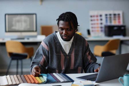 Téléchargez les photos : Designer afro-américain assis à son lieu de travail et en utilisant l'ordinateur tout en choisissant la couleur pour l'intérieur - en image libre de droit