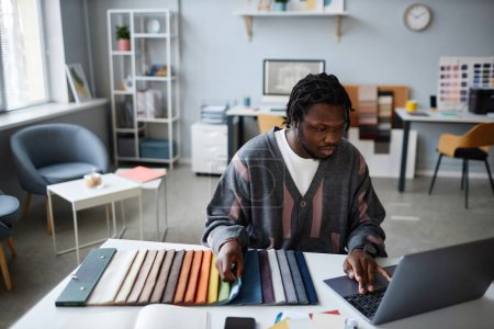 Téléchargez les photos : Designer afro-américain travaillant sur son lieu de travail avec le projet, il utilise un ordinateur pour adapter la couleur pour l'intérieur - en image libre de droit