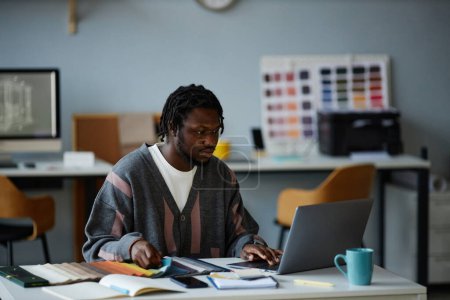 Téléchargez les photos : Concepteur afro-américain travaillant sur un ordinateur portable avec projet en ligne tout en étant assis sur son lieu de travail au bureau - en image libre de droit