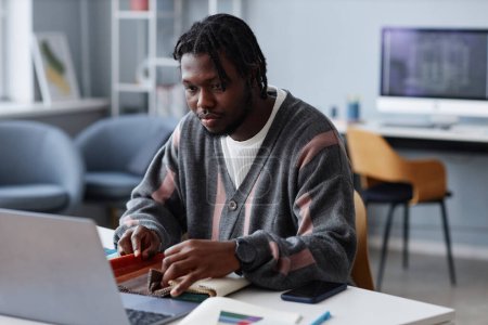 Téléchargez les photos : Jeune designer afro-américain travaillant sur un ordinateur portable et avec des échantillons de tissu assis sur son lieu de travail au bureau - en image libre de droit