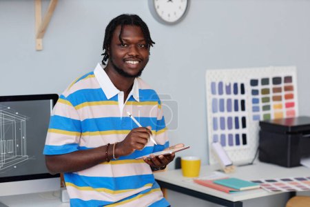 Téléchargez les photos : Portrait de designer afro-américain souriant à la caméra tout en prenant des notes sur tablette pc debout dans le bureau - en image libre de droit