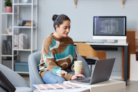 Téléchargez les photos : Jeune graphiste travaillant sur ordinateur portable avec projet et boire du café tout en étant assis à la table dans le bureau - en image libre de droit