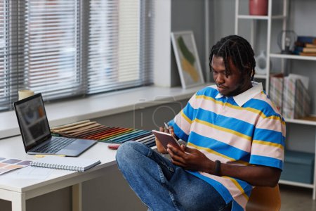 Téléchargez les photos : Projet de dessin de jeune designer afro-américain sur tablette PC assis sur son lieu de travail au bureau - en image libre de droit