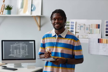 Téléchargez les photos : Portrait de designer afro-américain souriant à la caméra tout en prenant des notes dans un bloc-notes debout dans le bureau - en image libre de droit