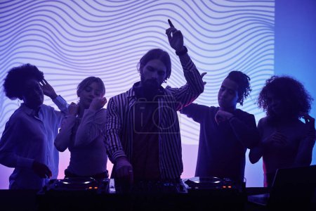 Foto de Foto retroiluminada de diversos grupos de jóvenes bailando con DJ en luces de neón en la fiesta retro disco - Imagen libre de derechos
