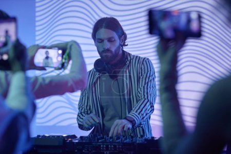 Foto de Retrato de DJ de pelo largo por estación de música en luces de neón entreteniendo a la multitud en la fiesta disco - Imagen libre de derechos