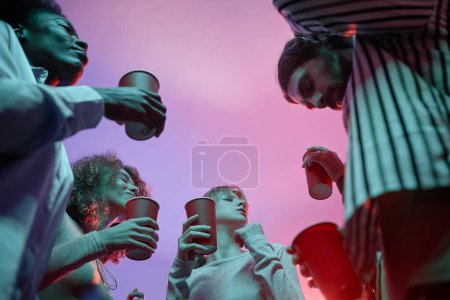 Foto de Vista de bajo ángulo en grupo de jóvenes en la fiesta de la casa sosteniendo copas rojas con alcohol y bailando en luz de neón rosa - Imagen libre de derechos