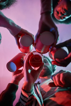 Téléchargez les photos : Vue à angle bas au groupe de jeunes tasses rouges cliquetis avec de l'alcool dans la lumière rose néon - en image libre de droit