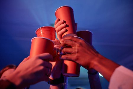 Foto de Primer plano de amigos que beben alcohol en la fiesta de la casa y tostadas con tazas de papel rojo en luz de neón - Imagen libre de derechos