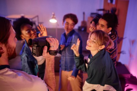 Foto de Grupo de jóvenes bailando en la fiesta de la casa con el hombre sosteniendo la cámara de vídeo retro y filmando video casero de amigos divirtiéndose, espacio para copiar - Imagen libre de derechos