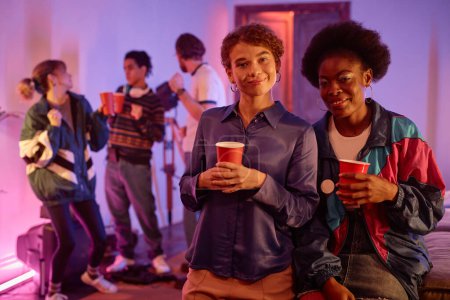 Foto de Retrato de dos chicas en la fiesta de la casa retro sosteniendo copas rojas y mirando a la cámara, espacio para copiar - Imagen libre de derechos
