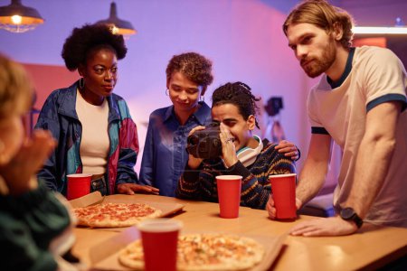 Téléchargez les photos : Portrait de jeune homme ethnique avec caméra vidéo rétro filmant des amis mangeant de la pizza à la fête de la maison - en image libre de droit