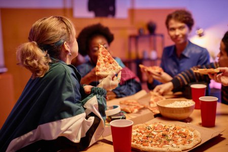 Photo for Side view portrait of young woman eating pizza with group of friends at house party 80s style, copy space - Royalty Free Image