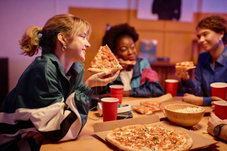 Photo for Side view portrait of smiling girl eating pizza with group of friends at house party 80s style, copy space - Royalty Free Image
