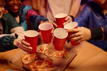 Foto de Primer plano de amigos sosteniendo tazas rojas y tostadas sobre la mesa con pizza en la fiesta de la casa, espacio para copiar - Imagen libre de derechos
