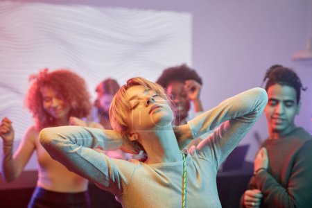 Foto de Retrato de mujer joven bailando con los ojos cerrados en discoteca y disfrutando de mucis con multitud en el fondo - Imagen libre de derechos