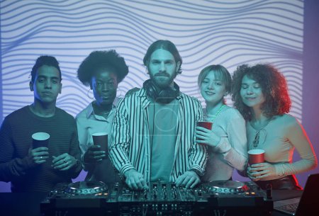 Téléchargez les photos : Portrait taille haute de DJ aux cheveux longs avec un groupe diversifié de personnes prenant des photos dans une boîte de nuit éclairée par néon - en image libre de droit