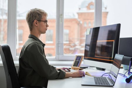 Téléchargez les photos : Vue latérale du jeune programmeur assis sur son lieu de travail et travaillant avec des codes de sécurité pour le programme à l'aide d'un ordinateur et d'un smartphone - en image libre de droit