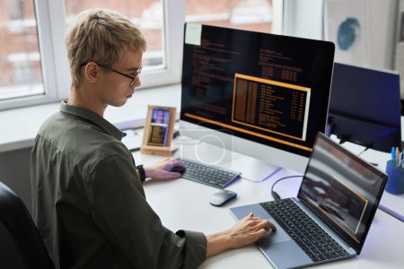 Foto de Vista trasera del joven programador en anteojos escribiendo códigos en diferentes dispositivos mientras trabaja en su lugar de trabajo en la oficina de TI - Imagen libre de derechos